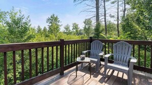 A wooden deck with two Adirondack chairs and a small table, overlooking a forested area. - A-Frame Hot tub Retreat - Firepit, Grill, EV & Dog Friendly, Close to N. Conway. (Fryeburg)