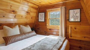 A cozy wooden cabin bedroom with a bed, pillows, a window, and framed artwork on the walls. - A-Frame Hot tub Retreat - Firepit, Grill, EV & Dog Friendly, Close to N. Conway. (Fryeburg)