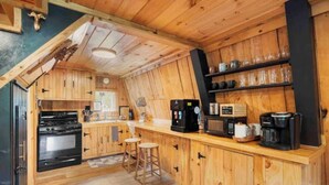 A kitchen with wooden cabinets, a stove, stools, and a coffee station. - A-Frame Hot tub Retreat - Firepit, Grill, EV & Dog Friendly, Close to N. Conway. (Fryeburg)