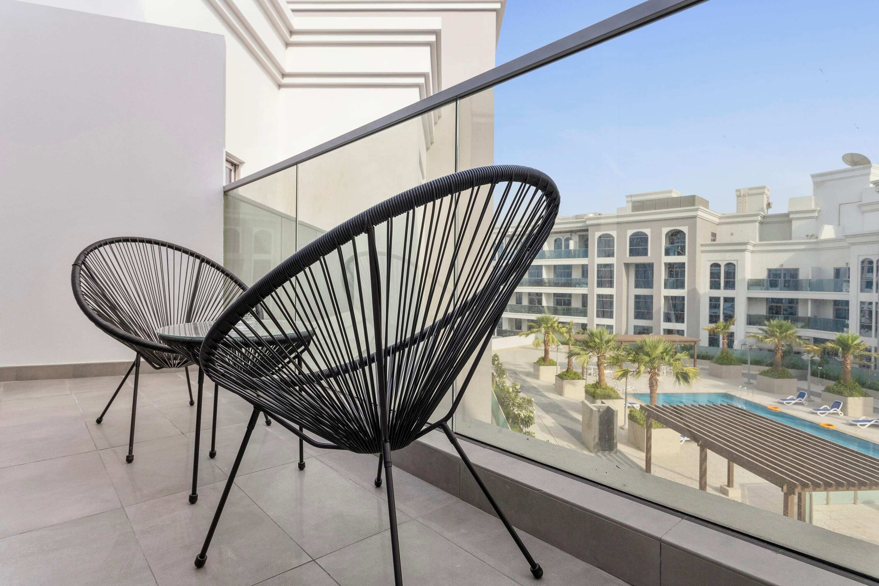A balcony with two black wire-framed chairs, a tiled floor, and a view of a pool and buildings.