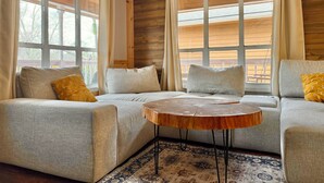 Una habitación acogedora con sofá, una mesa redonda de madera y ventanas grandes con cortinas.
