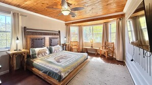 Un dormitorio con techo de madera, cama con colcha estampada, ventilador de techo y una ventana grande con cortinas.