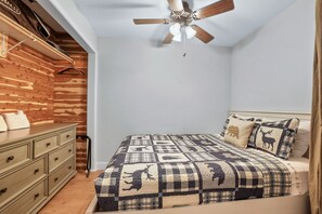 Un dormitorio con pared de madera, cama con manta acolchada, mesita de noche y ventilador de techo.