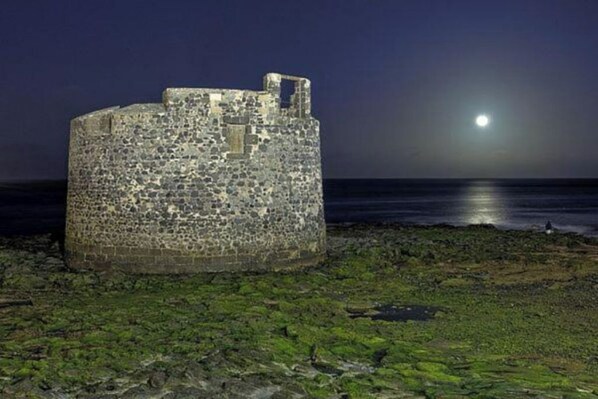 Una estructura de piedra con techo parcialmente conservado, ubicada en una orilla rocosa con vista al mar y una luna llena de fondo.