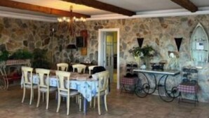A rustic dining area with stone walls, wooden beams, and a tiled floor. There are several white chairs around a table with a blue tablecloth. A bicycle is parked against the wall, and a small table with a checkered tablecloth is visible.
