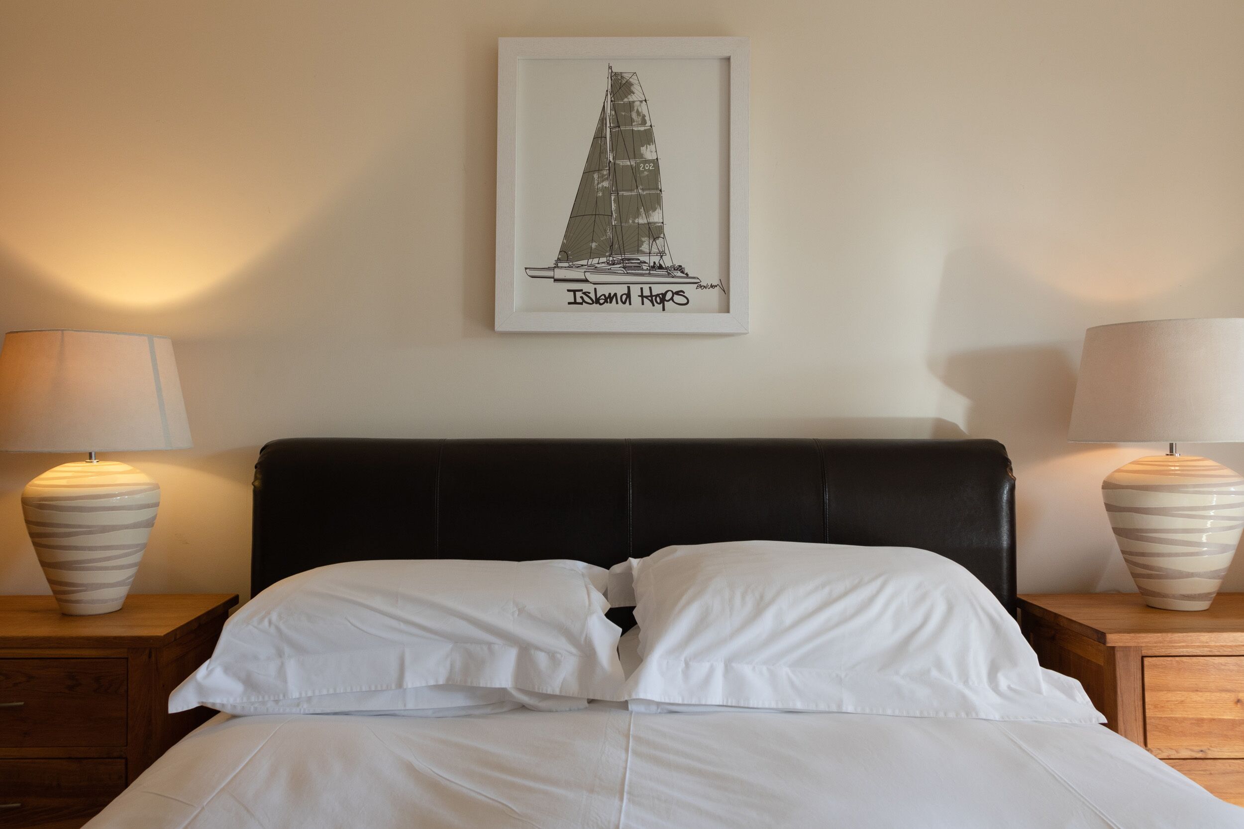 A neatly made bed with white linens, a black headboard, and two bedside lamps. A framed picture of a sailboat labeled "Island Hops" hangs on the wall above the bed.