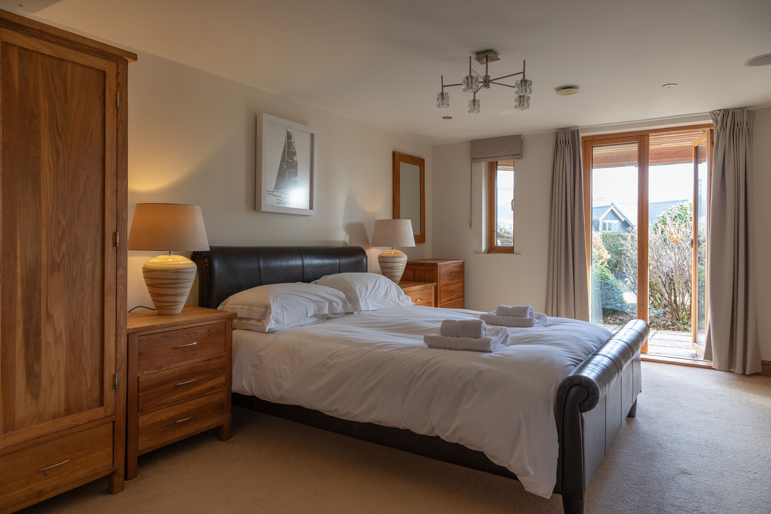 A bedroom with a large bed, wooden wardrobe, two bedside tables with lamps, a ceiling light fixture, and a view of the outdoors through a sliding glass door.