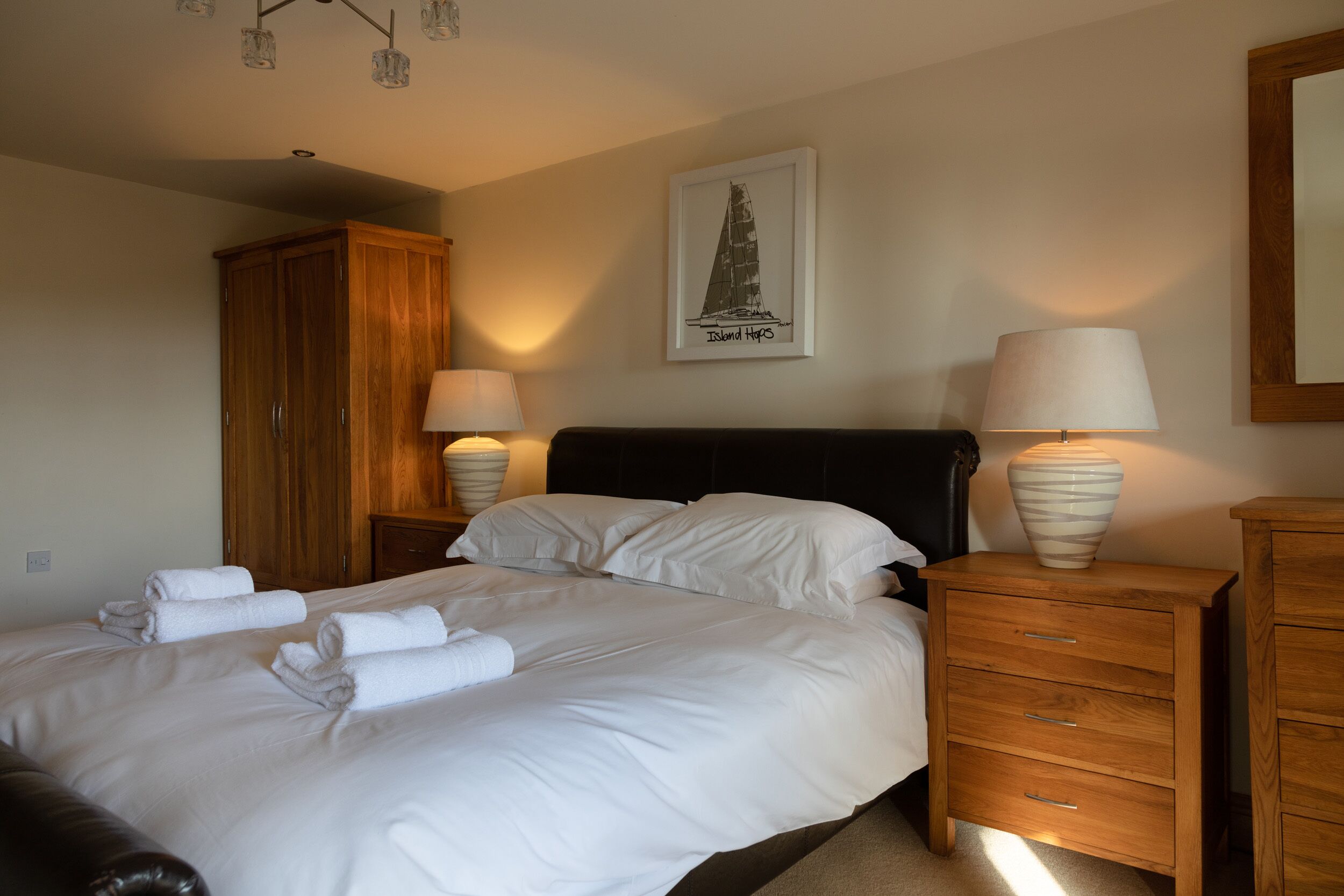 A neatly made bed with white linens and rolled towels, a wooden wardrobe, a framed picture on the wall, and two bedside tables with lamps.