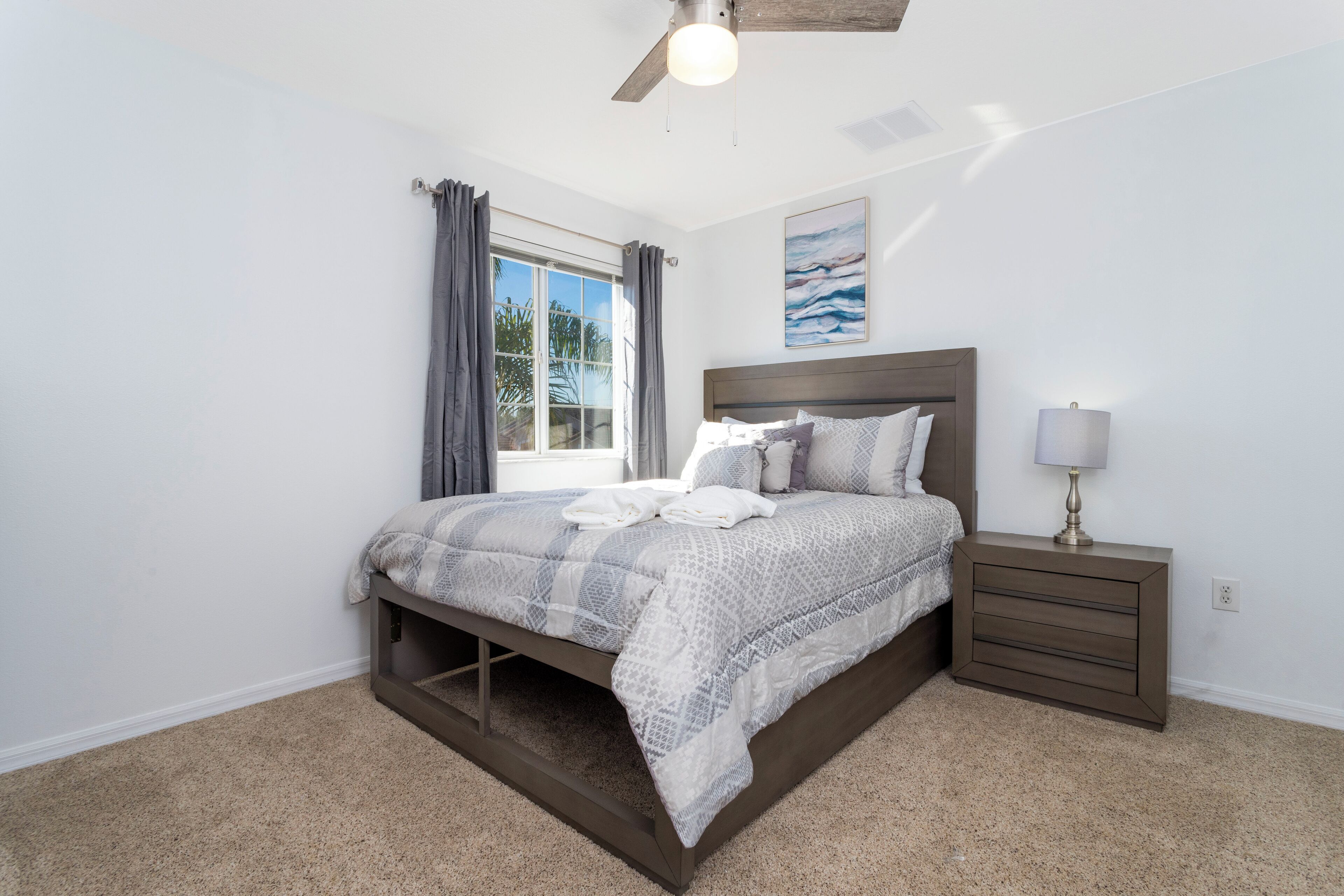 A bedroom with a bed, a nightstand, a lamp, a ceiling fan, and a window with curtains.