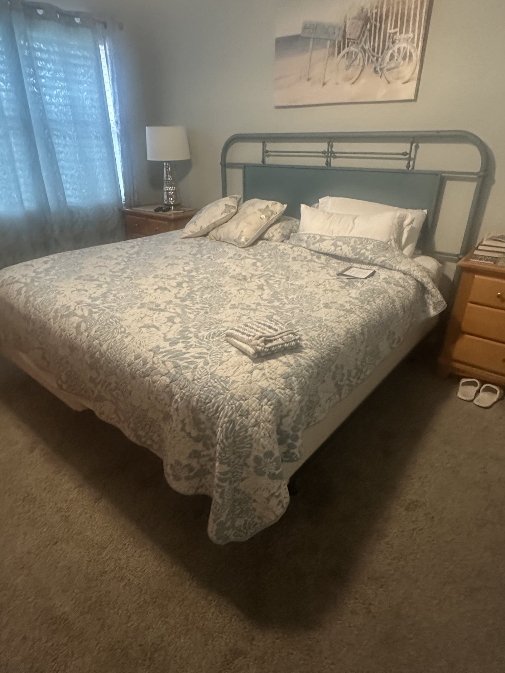 A bed with a patterned bedspread, pillows, a nightstand with a lamp, and a pair of slippers on the floor.