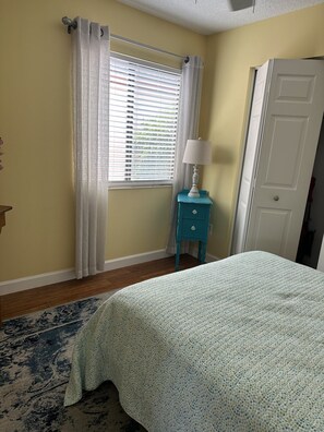 A bedroom with a bed, a nightstand, a window with blinds, a door, and a patterned rug. - Casa Bella at Sea Place (St. Augustine)