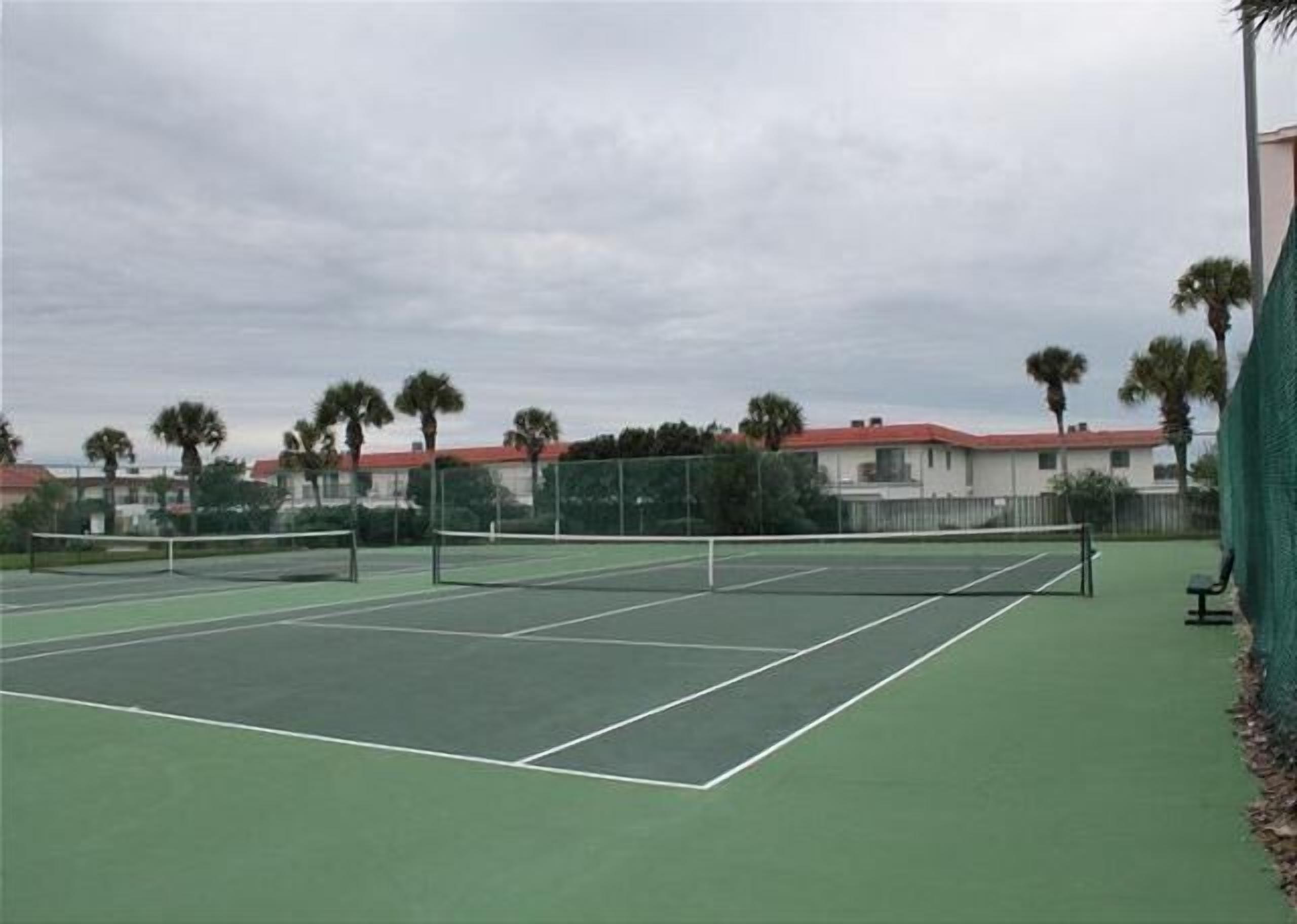 Un court de tennis en plein air, doté d’un filet, entouré d’une clôture et bordé de palmiers.