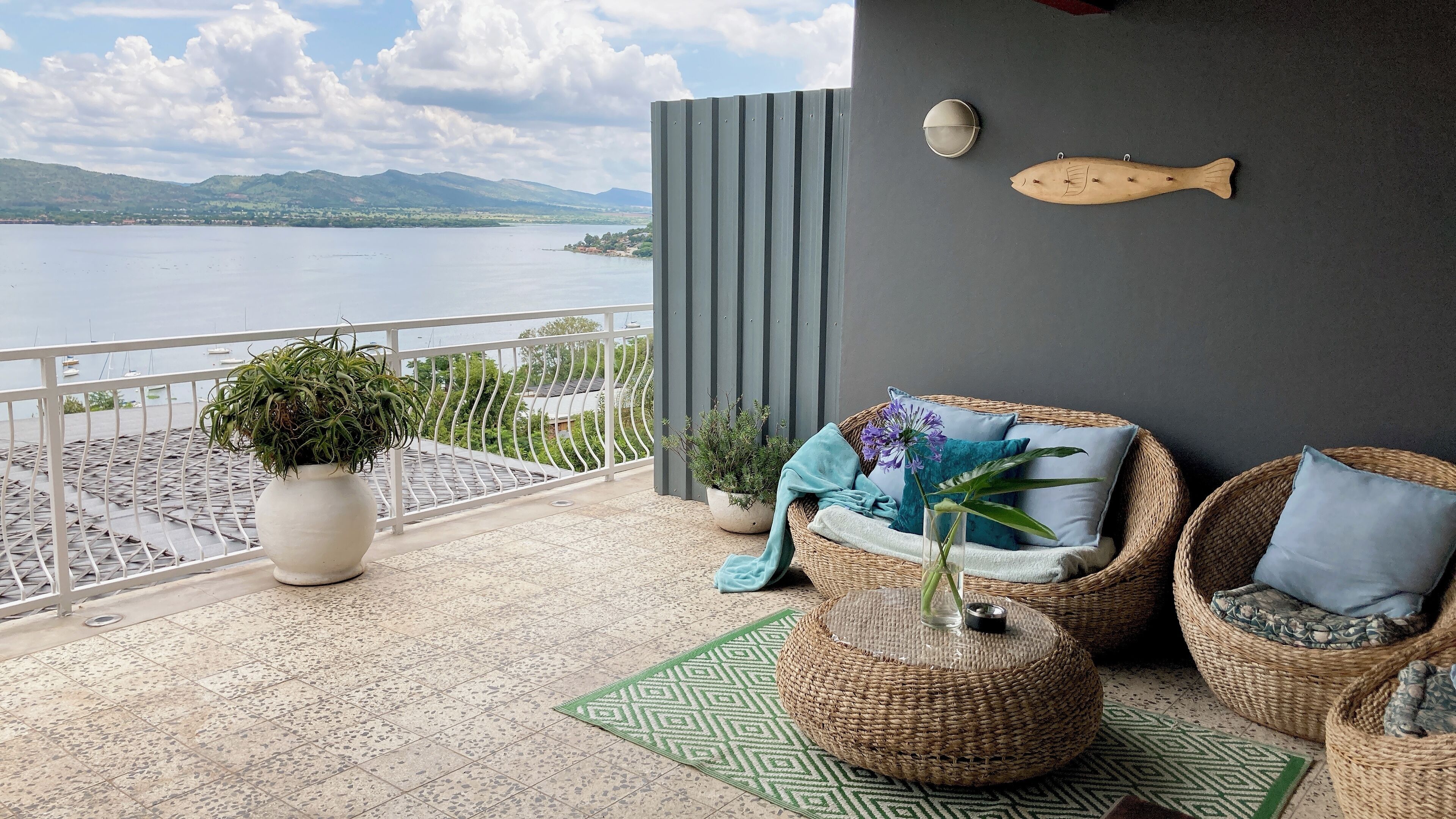 A balcony with wicker furniture, potted plants, and a view of a lake and mountains.