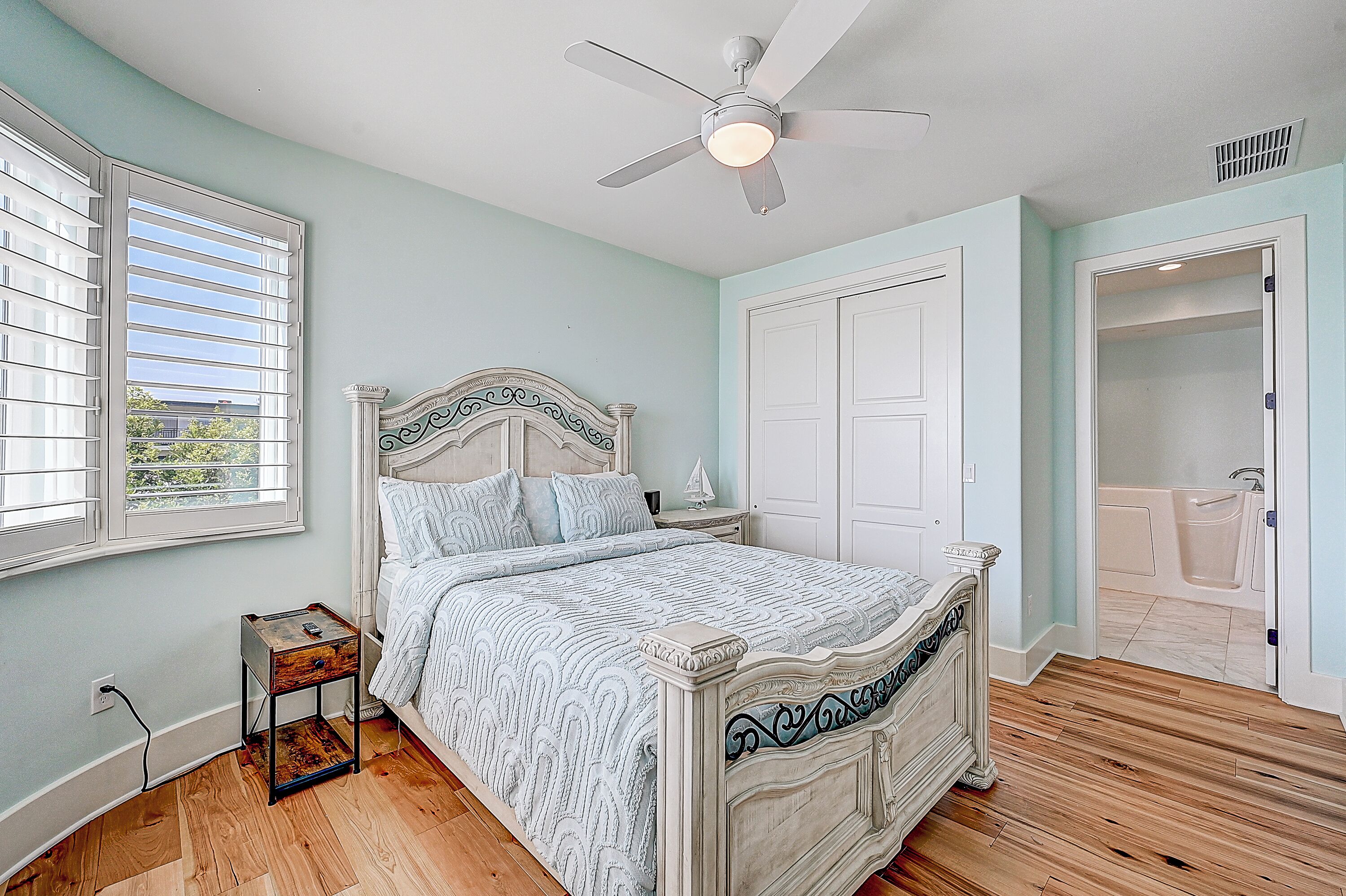 A bedroom with a bed, a nightstand, a ceiling fan, a window with blinds, and a door leading to a bathroom.