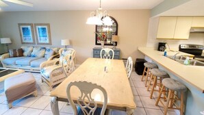 Un comedor con una mesa de madera y sillas blancas, un sofá con cojines, y una zona de cocina con un lavamanos y taburetes.