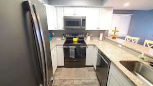 A compact kitchen with white cabinets, a stainless steel refrigerator, a microwave, an oven, and a dishwasher. - Ocean Villas 6 (St. Augustine)