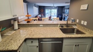 A kitchen with granite countertops, a stainless steel sink, and a dishwasher. There is a dining area with a table and chairs visible through the window. - Ocean Villas 6 (St. Augustine)