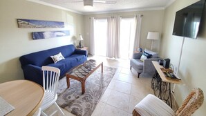 A living room with a blue sofa, a coffee table, a TV, and a ceiling fan. - Pelican Inlet A206 (St. Augustine)