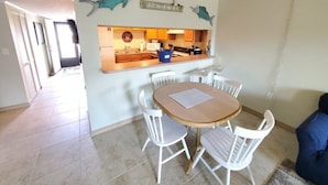 A dining area with a wooden table and white chairs, a kitchen with wooden cabinets, and a wall with shark decorations and a sign. - Pelican Inlet A206 (St. Augustine)