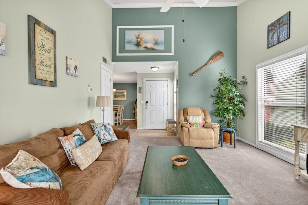 A living room with a brown couch, a green coffee table, and a beige armchair. There is a door leading to another room, a plant, and a window with blinds.