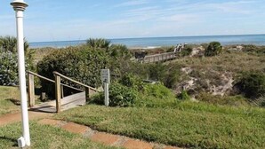 Ein Küstenpfad führt zu einem Strand mit einer Holztreppe und einem weißen Pfosten.
