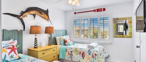 A bedroom with two beds, a ceiling fan, a window with blinds, and a mounted fish tail decoration.