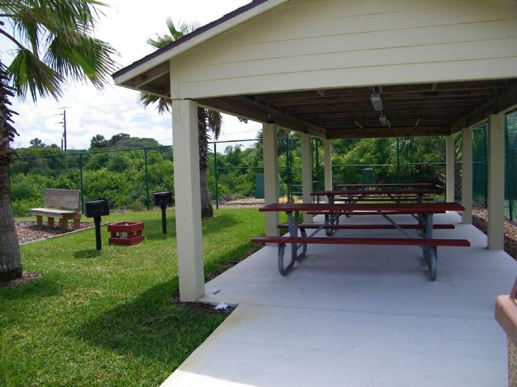 A covered picnic area with benches and a table, surrounded by greenery and a palm tree.