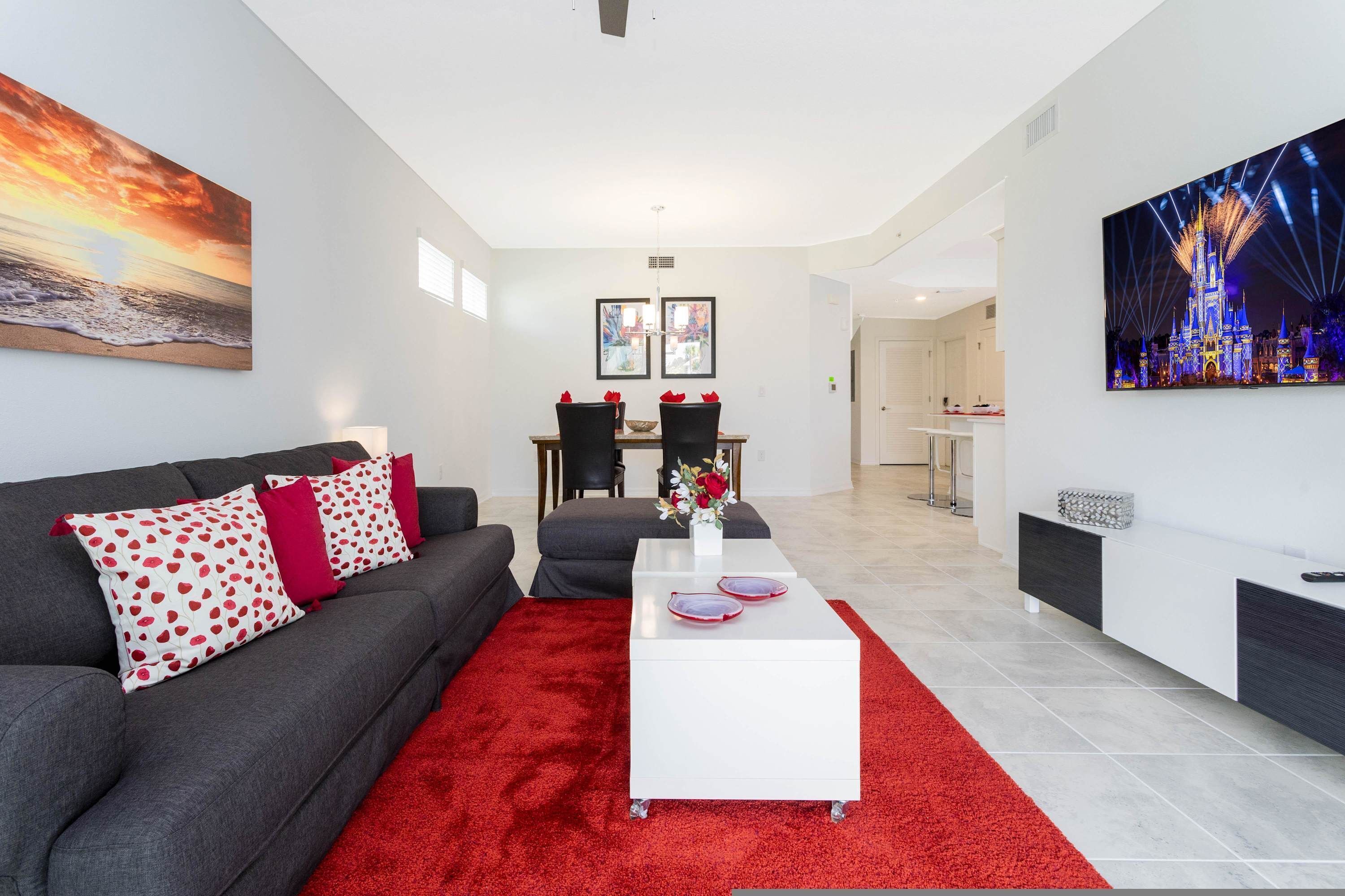 Una sala de estar moderna con un sofá gris, almohadones blancos y rojos con estampado, una mesa de centro blanca y una alfombra roja. Hay dos cuadros enmarcados en la pared, un área de comedor al fondo y un cuadro grande de un castillo.