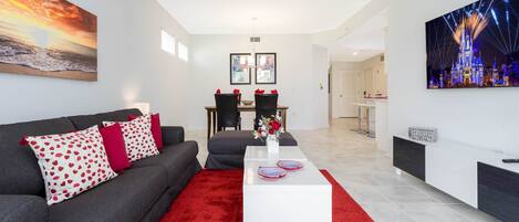 Una sala de estar moderna con un sofá gris, almohadones blancos y rojos con estampado, una mesa de centro blanca y una alfombra roja. Hay dos cuadros enmarcados en la pared, un área de comedor al fondo y un cuadro grande de un castillo.
