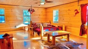 A log cabin interior with a wooden floor, a bed with a red blanket, a table, and a ceiling fan. - 5 min from Parkway. Cozy cabin! (Sevierville)