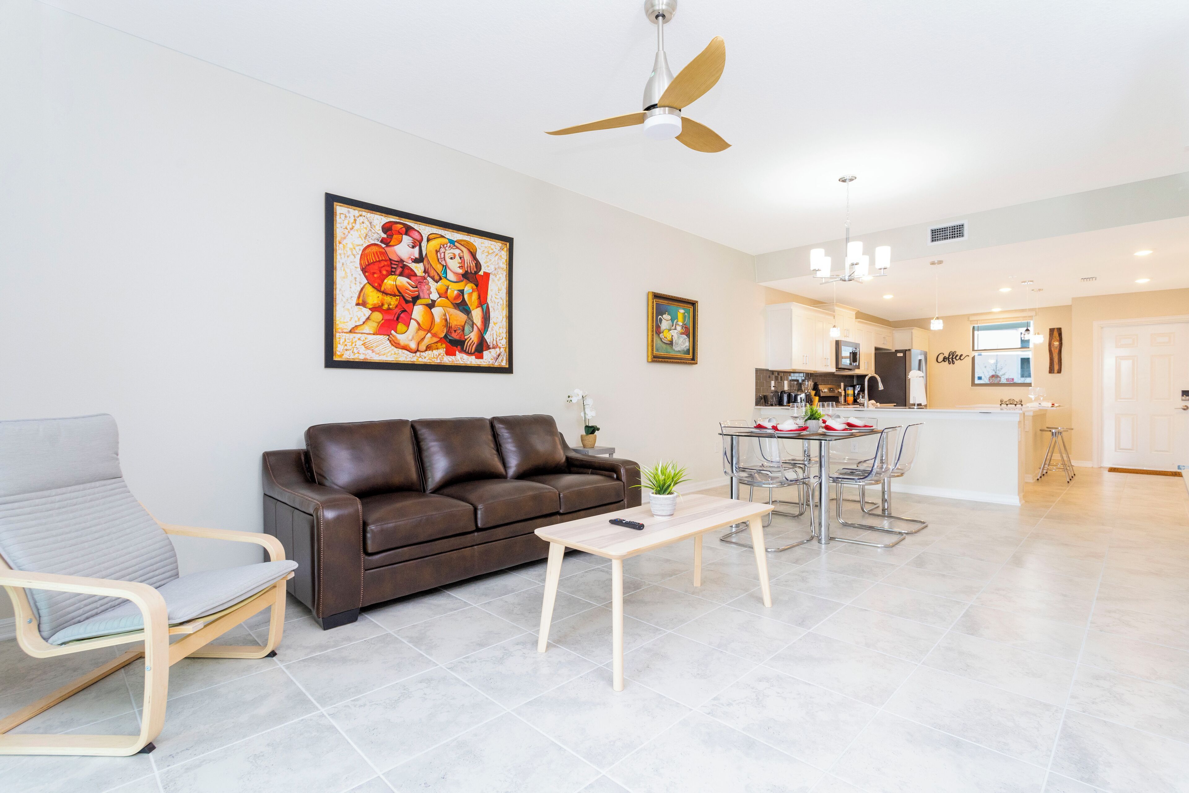 Een moderne woonkamer met een bruine leren bank, een grijze fauteuil en een houten salontafel. Een plafondventilator is zichtbaar, en er is een keuken in de achtergrond.