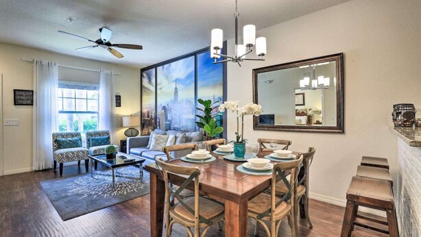 A dining area with a wooden table set for six, surrounded by wicker chairs. A ceiling fan is visible above the table, and a large window with curtains is in the background.