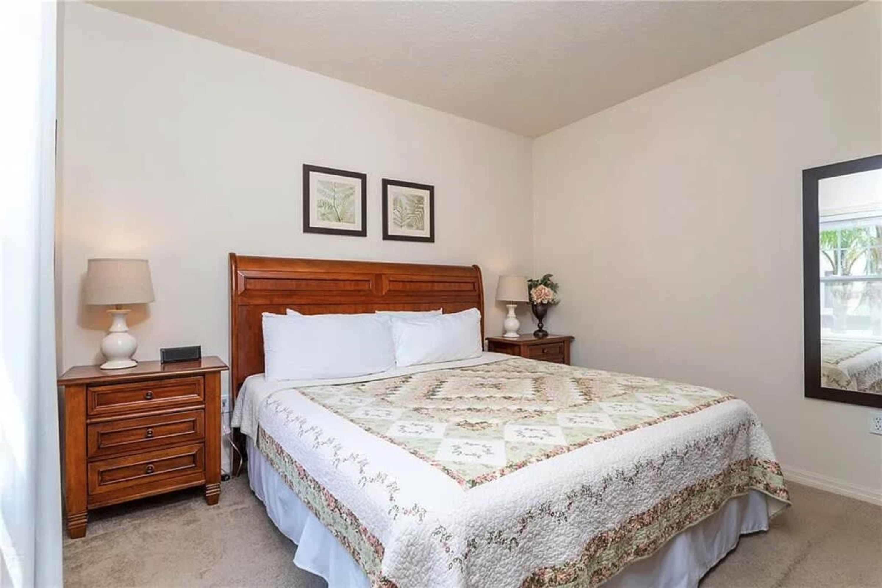 A bedroom with a wooden headboard, a floral quilt, a nightstand with a lamp, and two framed pictures on the wall.