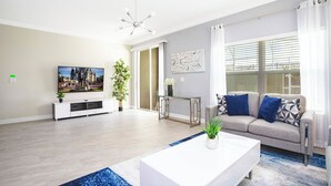 A modern living room with a flat-screen TV, a sofa with patterned cushions, a white coffee table, and a blue rug. - Stunning 5 Bd w Pool Close to Disney Champions Gate 1012 (Davenport)