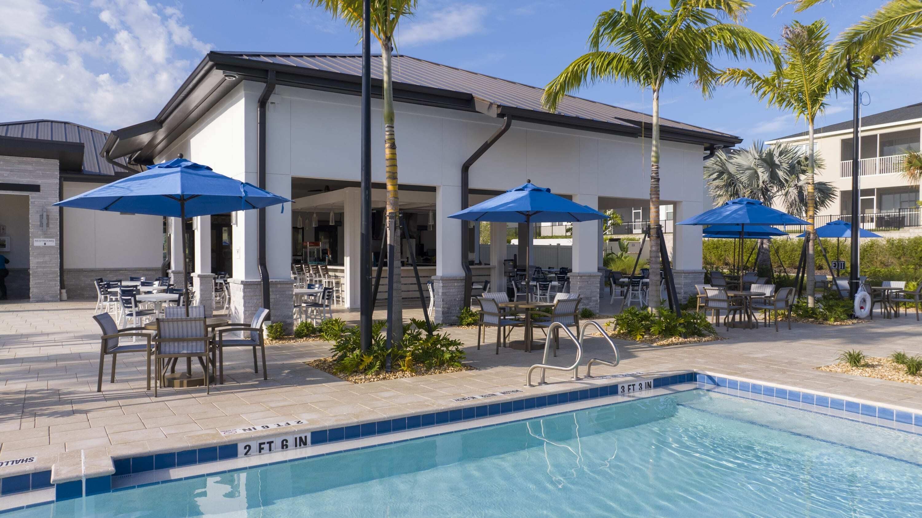 A poolside area with outdoor seating and umbrellas, a clear pool, and a building with a white facade and dark roof.