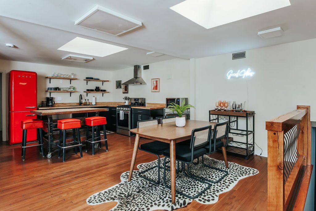 A modern kitchen with a red refrigerator, wooden bar stools, a dining table with chairs, and a neon sign on the wall.