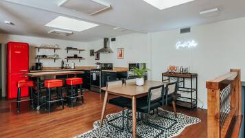 A modern kitchen with a red refrigerator, wooden bar stools, a dining table with chairs, and a neon sign on the wall.