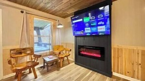 A cozy living room with a fireplace, wooden furniture, and a mounted flat-screen TV displaying various channels. - Cozy cabin! Great Location! (Sevierville)