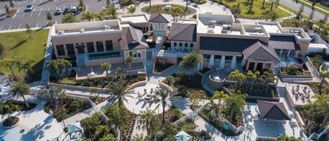 An aerial view of a hotel with multiple buildings, a central courtyard with palm trees, and a swimming pool area.