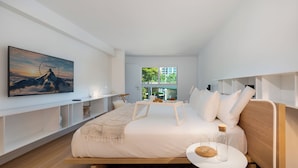 Een moderne slaapkamer met een bed, nachtkastje, een aan de muur bevestigde televisie en een raam met uitzicht op groen.