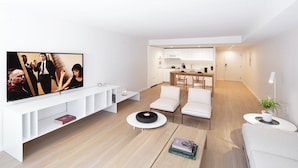 Een moderne woonkamer met een flatscreen televisie, witte banken, een salontafel en een keuken in de achtergrond.