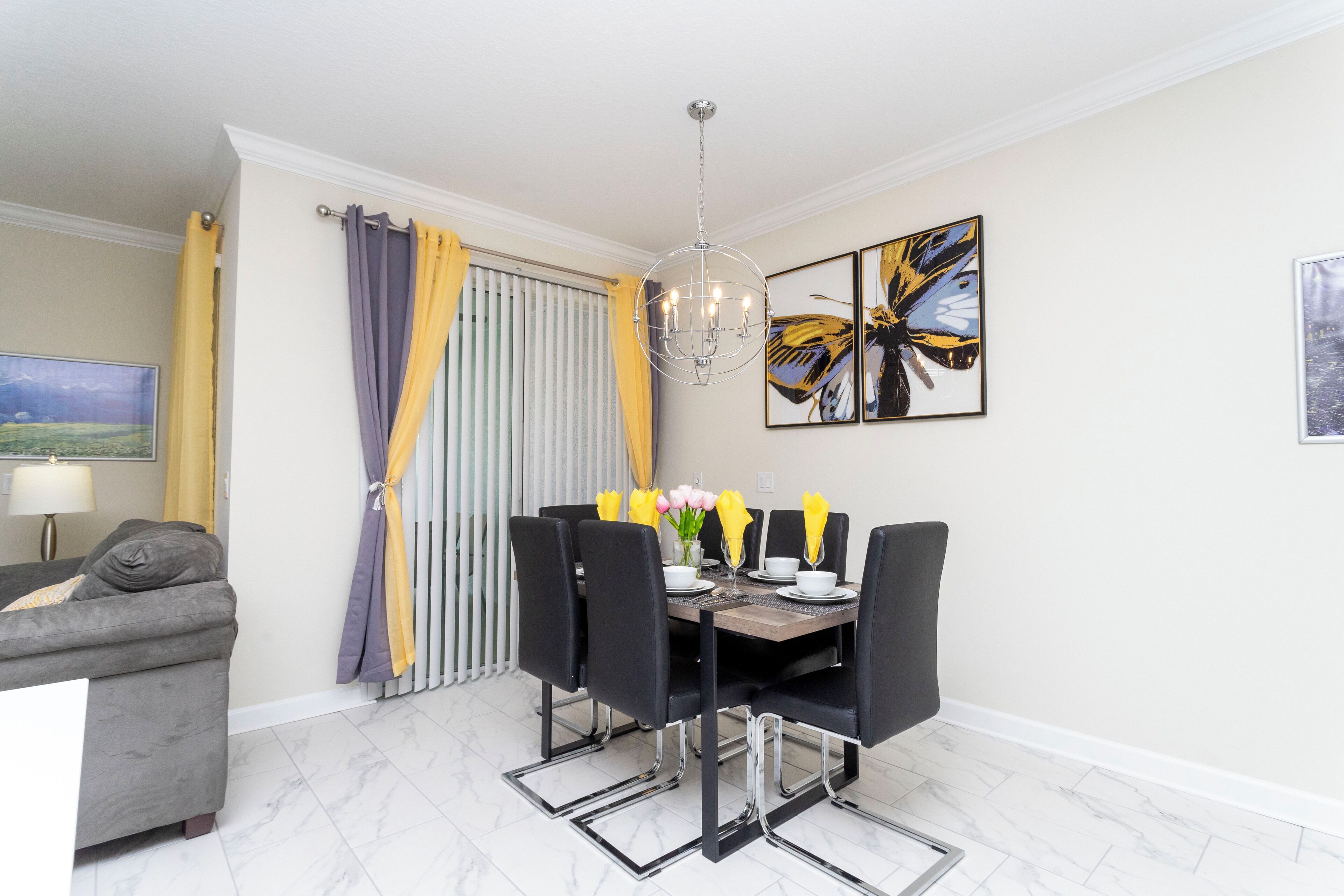 A dining area with a table set for six, surrounded by black chairs, a chandelier, and two butterfly paintings on the wall.