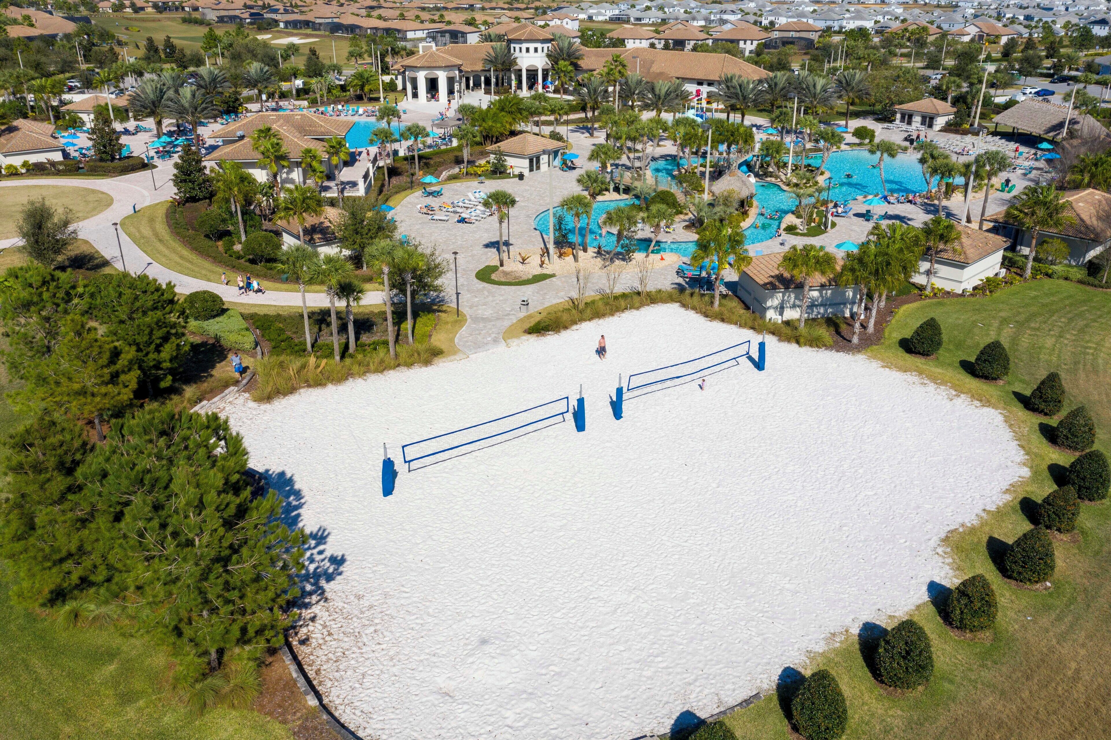 An aerial view of a resort with multiple swimming pools, palm trees, and a central open area with a white surface.
