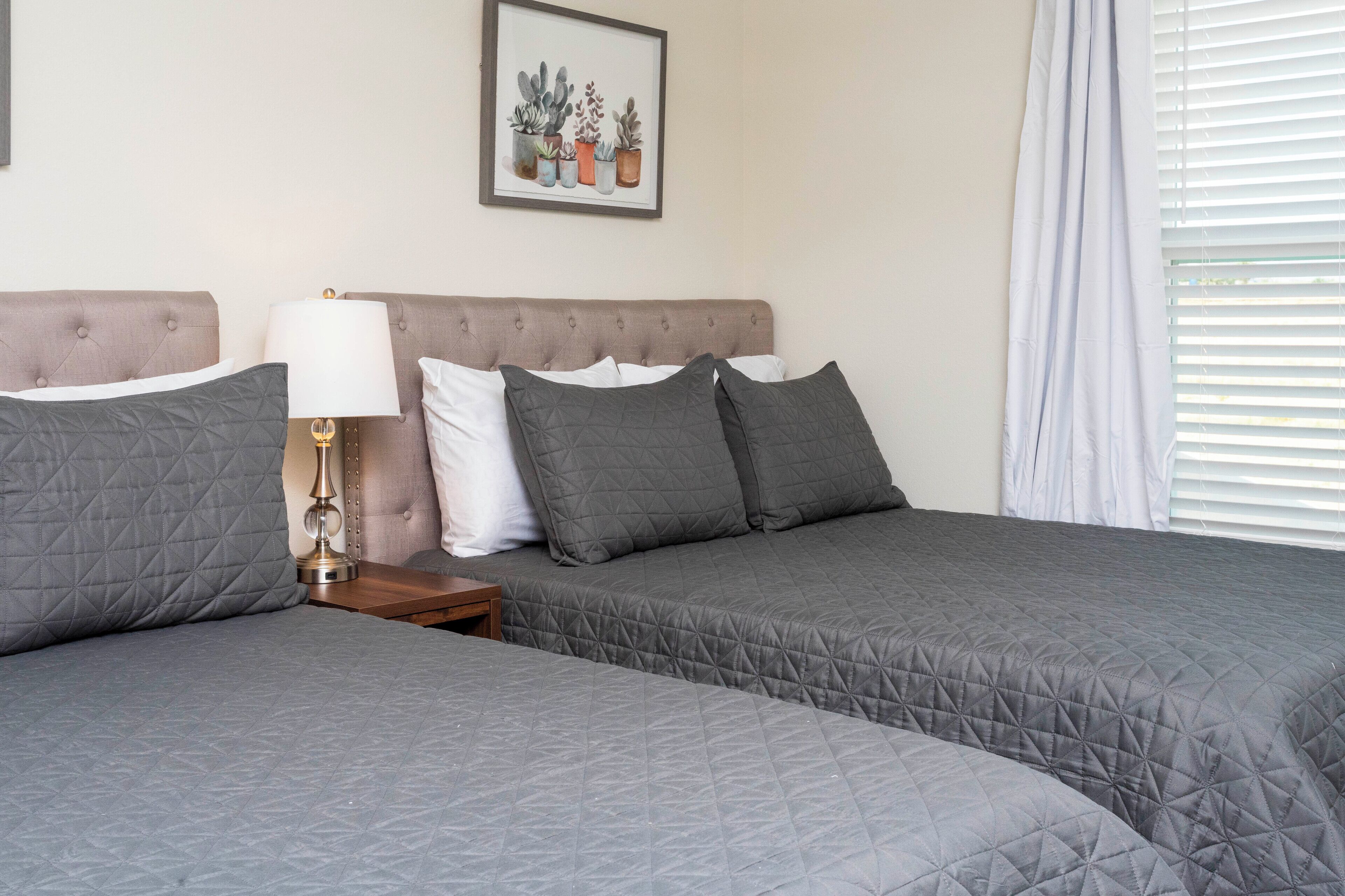 A hotel room with two beds, a nightstand, a lamp, and a framed picture of plants on the wall.