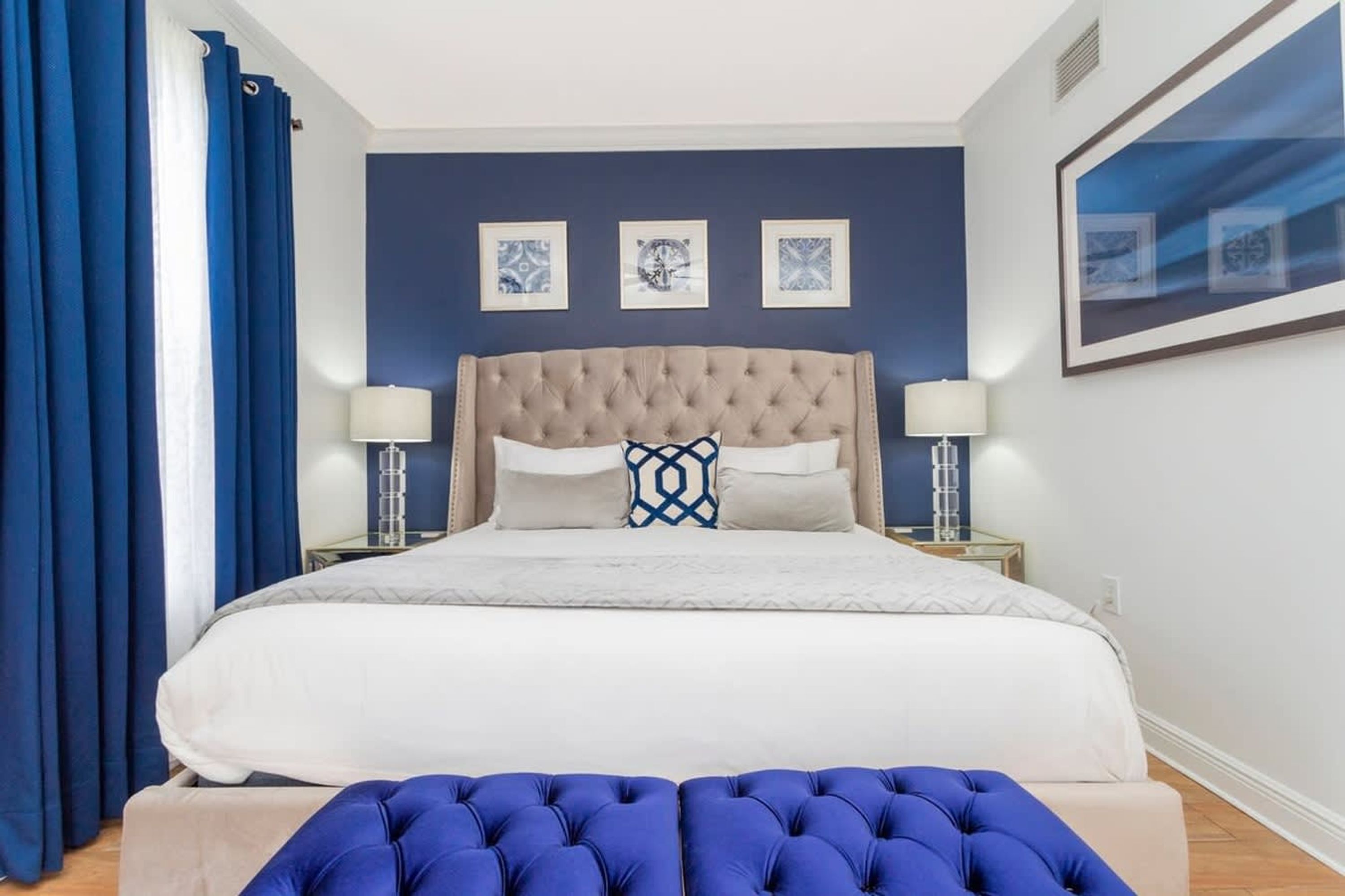 A bedroom with a large bed, two bedside tables, a blue upholstered bench, and three framed pictures on the wall.