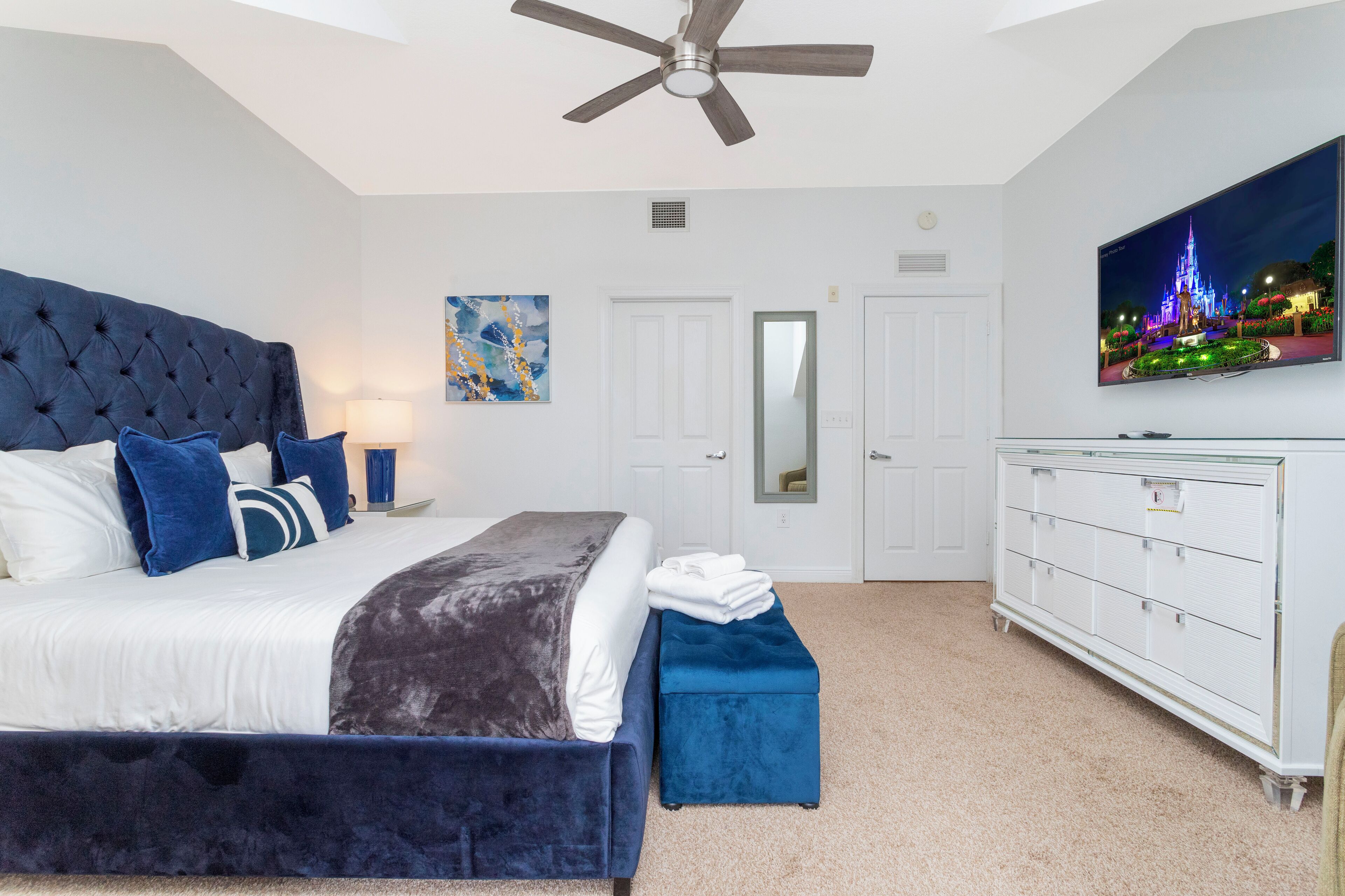 A bedroom with a large bed, a ceiling fan, a TV on the wall, and a dresser.