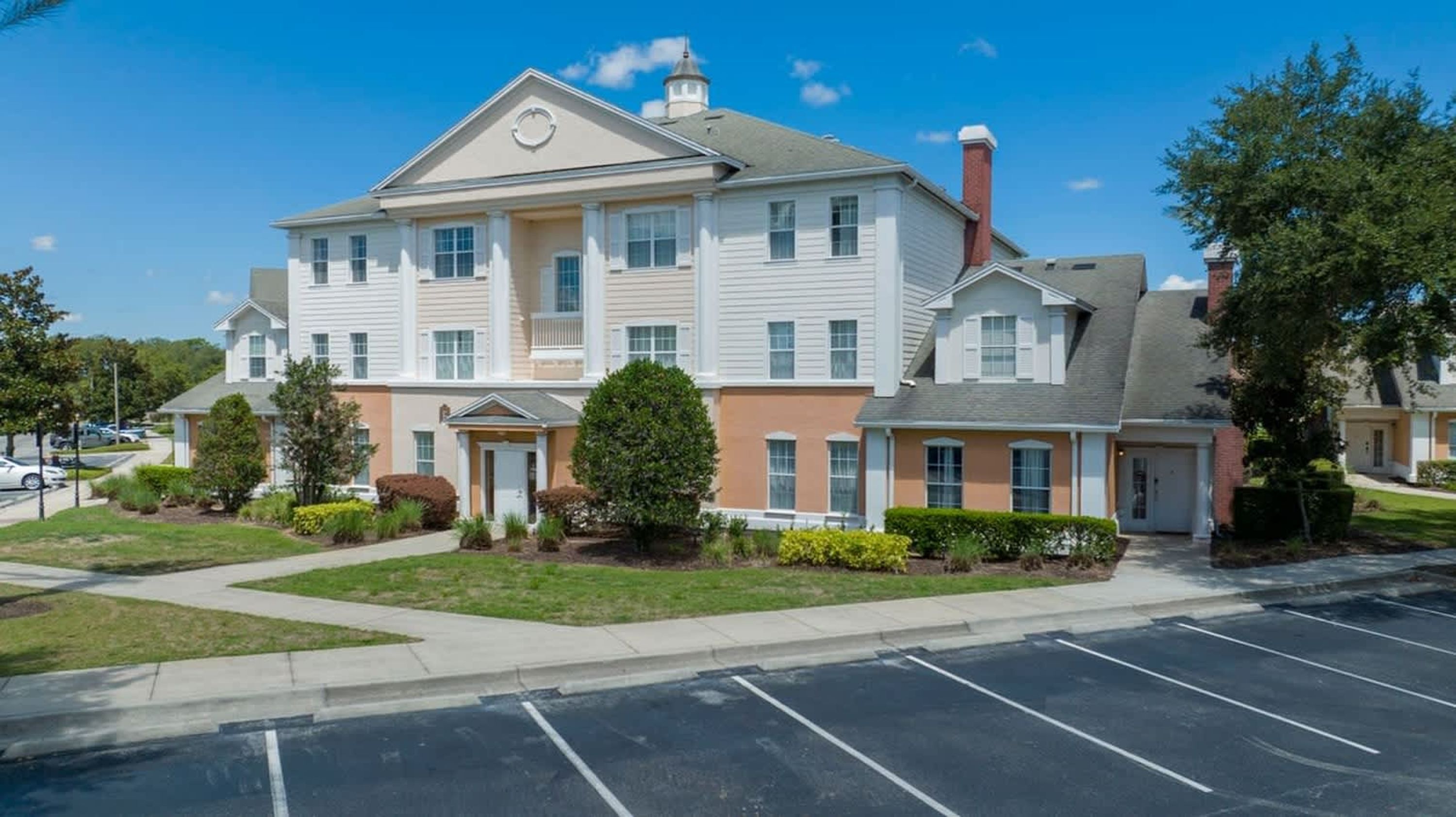 A multi-story residential building with a central entrance, a parking lot, and a clear sky.
