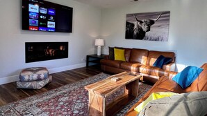 Una sala de estar con un televisor de pantalla plana montado en la pared que muestra varios servicios de transmisión, un sofá de cuero marrón con cojines coloridos, una mesa de centro de madera, un tapete estampado y una obra de arte en la pared con un toro.