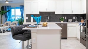 A modern kitchen with white cabinets, a black backsplash, and stainless steel appliances. There is a dining area with a black bar stool and a table set for two. - Luxurious 3Bd 8 Guests Storey Lake Resort Close to Disney 201 3131 (Kissimmee)