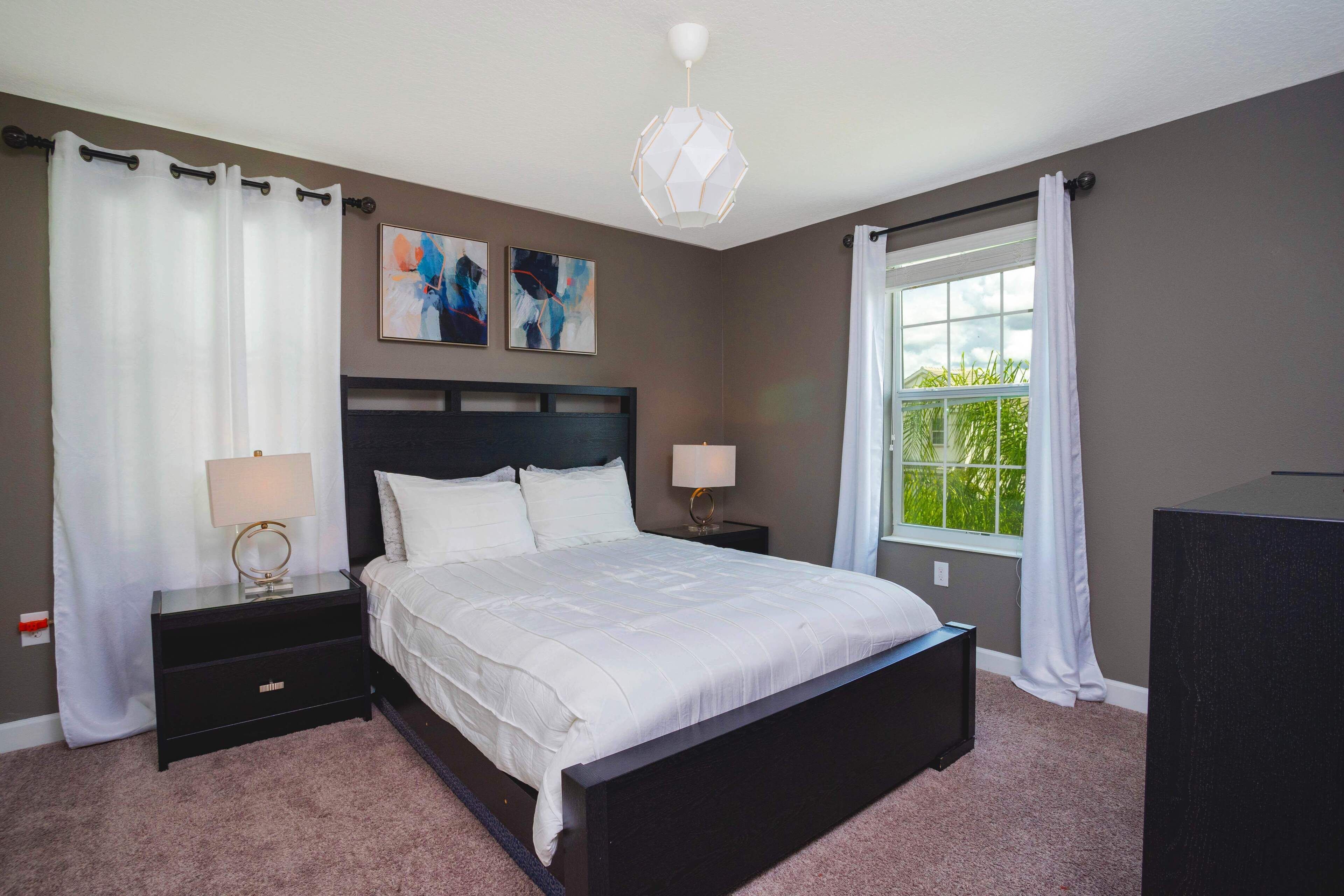 A bedroom with a bed, two bedside tables, a nightstand, a window with curtains, and abstract wall art.